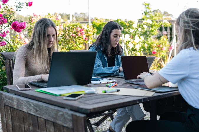Three university college students studying in the garden at home preparing for exams, taking notes.