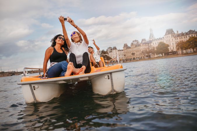Friends sitting in front pedal boat and taking selfie with man in background
