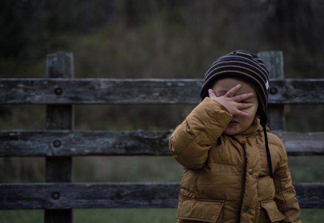 Toddler cover his face standing near wooden fence