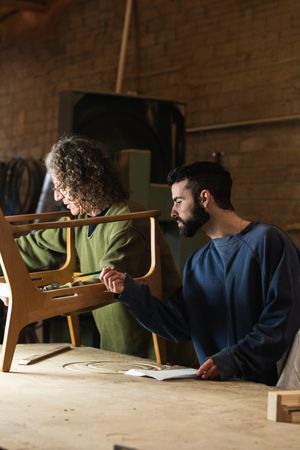 Craftsman creating wooden chair with coworker drawing sketches