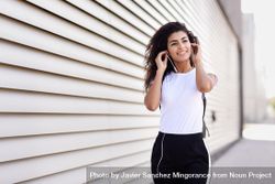 Arab woman in sport clothes with curly hair standing in front of wall adjusting headphones 5rge14