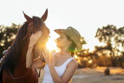Young woman in hat walking with her horse in the countryside at sunset