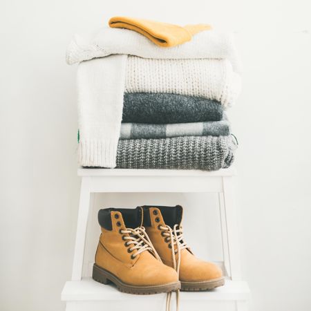 Pile of clean, folded sweaters on light background with yellow boots, square crop