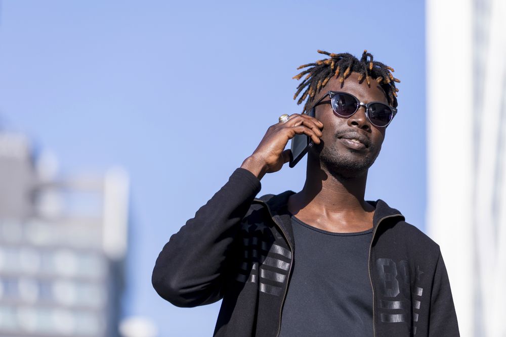Serious Black male with sunglasses standing against cityscape on the street while using a mobile phone in sunny day