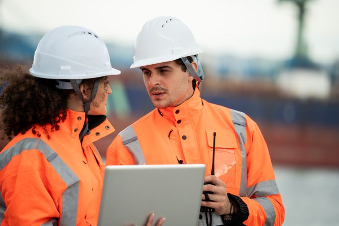 Workers in safety helmets examine laptop screen at sea, Freight ship in background, Dockyard engineers collaborate by harbor, man uses walkie-talkie, Port facility teamwork, Logistics industrial