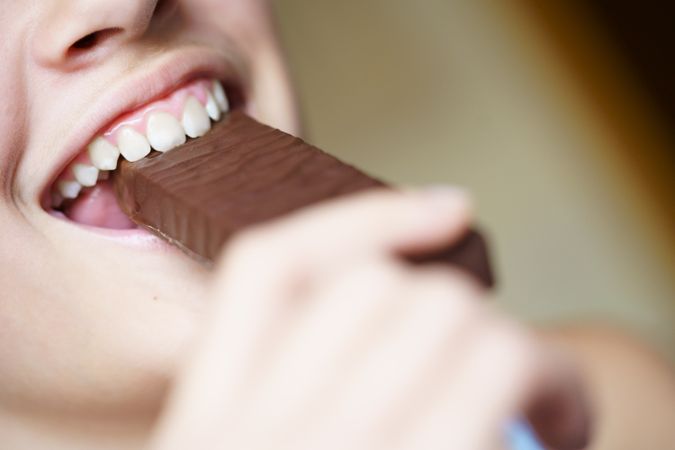Happy crop anonymous teenage girl taking bite of chocolate bar