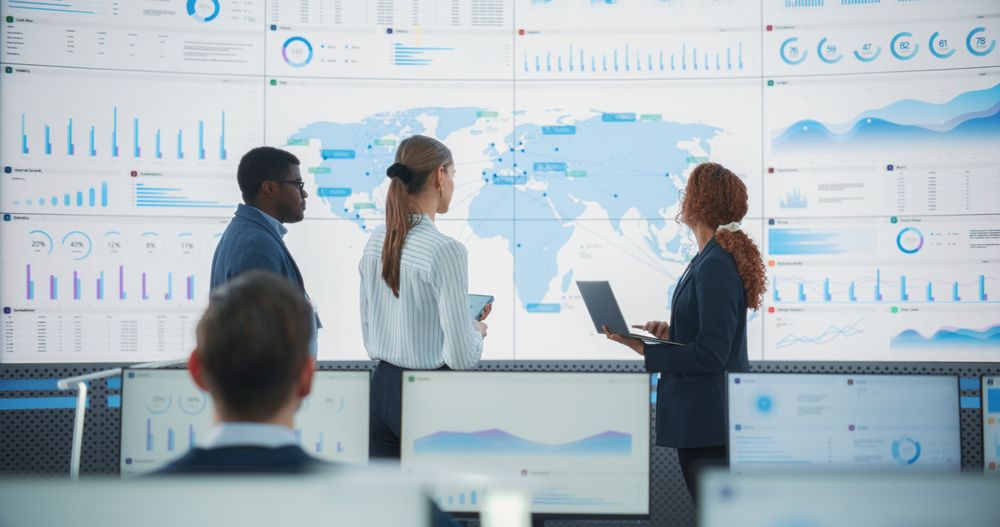 Diverse Team Of Data Science Department Discussing New Markets In Front of Big Digital Screen With World Map In Monitoring Office.