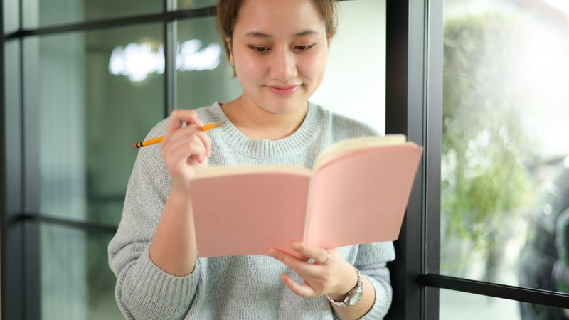 A young woman standing by the window reading a book.