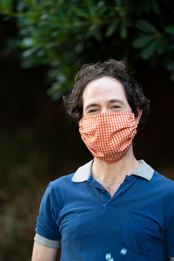 Close up portrait of man in PPE mask smiling and looking at camera in front of green bushes