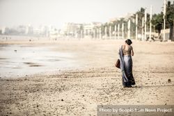 Woman in gray sari and holding a suitcase walking on brown sand 0Vx834
