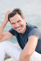 Confident adult man smiling at camera sitting on rocky beach