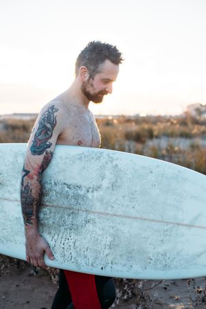 Tattooed man looking down with surfboard under his arm