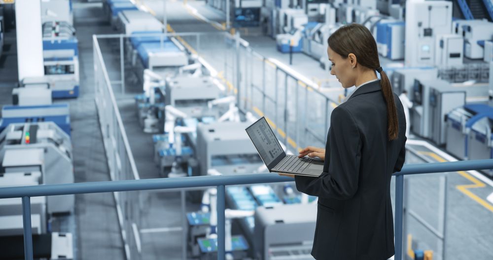 Portrait of a White Female Engineer Using Laptop Computer and Looking Around a Factory Facility with Robotic Arms Producing Modern Electronic Components for Tech Industries