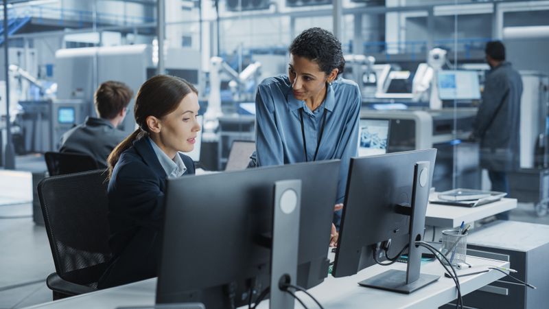 White Female Engineer And Hispanic Product Manager Using Desktop Computer And Talking At Automated Factory With Robotic Hands.