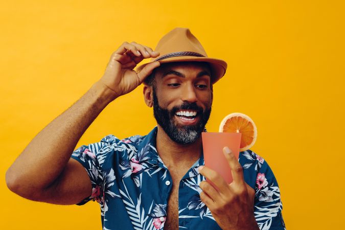 Content male adjusting hat while wearing colorful shirt and holding cocktail with yellow background
