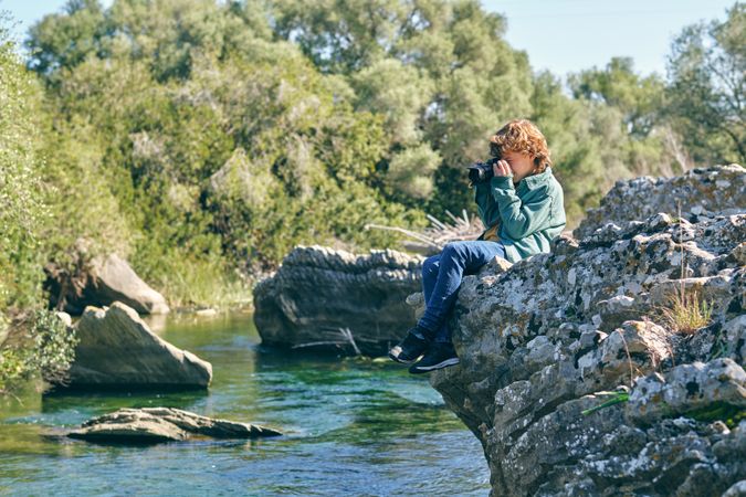 Curious boy taking photo while sitting on rocky coast near lake