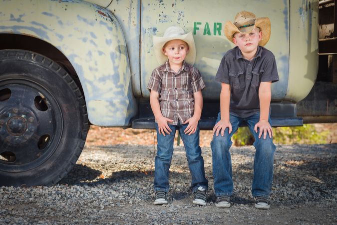 Young Boys Wearing Cowboy Hats Leaning Against an Antique Truck in a Rustic Setting