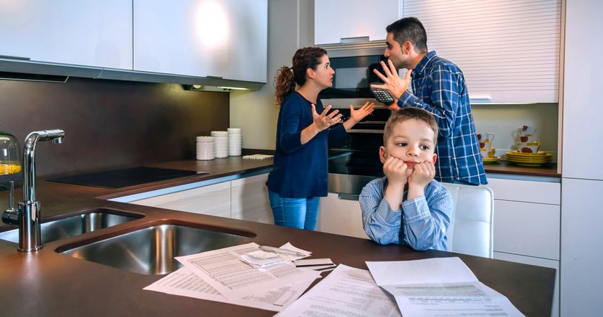 Couple fighting over money in front of son with bills strewn on the kitchen counter
