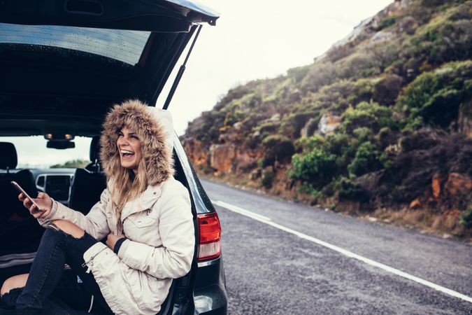 Smiling woman sitting in car trunk with a smartphone