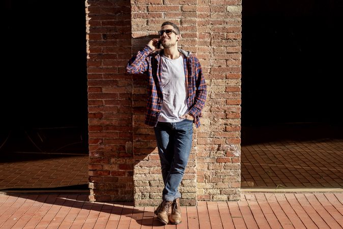 Man leaning back on a brick wall wearing sunglasses and chatting on smartphone outdoors