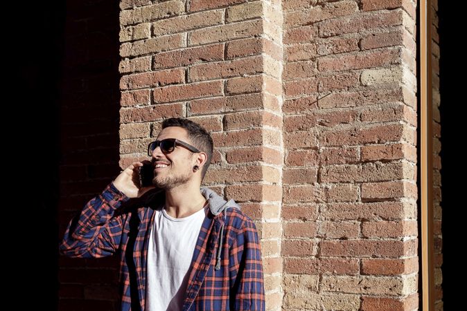 Bearded man leaning on a brick wall wearing sunglasses while using a smartphone