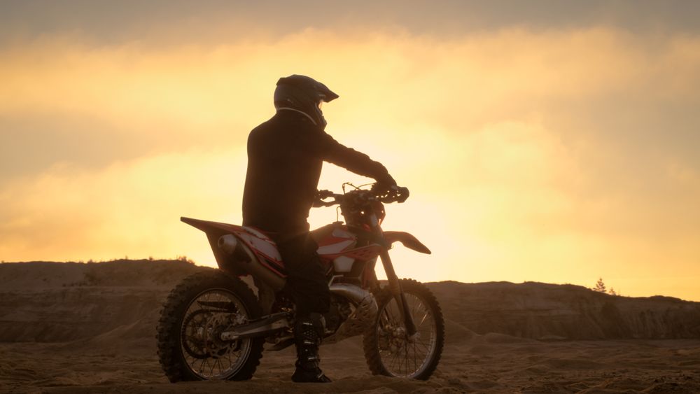 Professional FMX Motorcycle Rider Rests on His Bike and Overlooks Hard Sandy Off-Road Terrain.