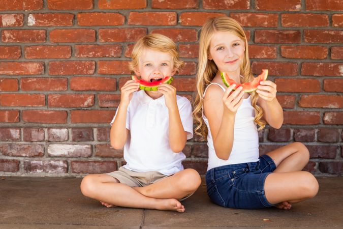 Cute Young Boy and Girl Eating Watermelon Against Brick Wall