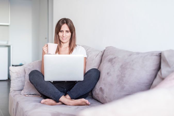 Smiling woman sitting on sofa with a cup of coffee working on her laptop in the morning