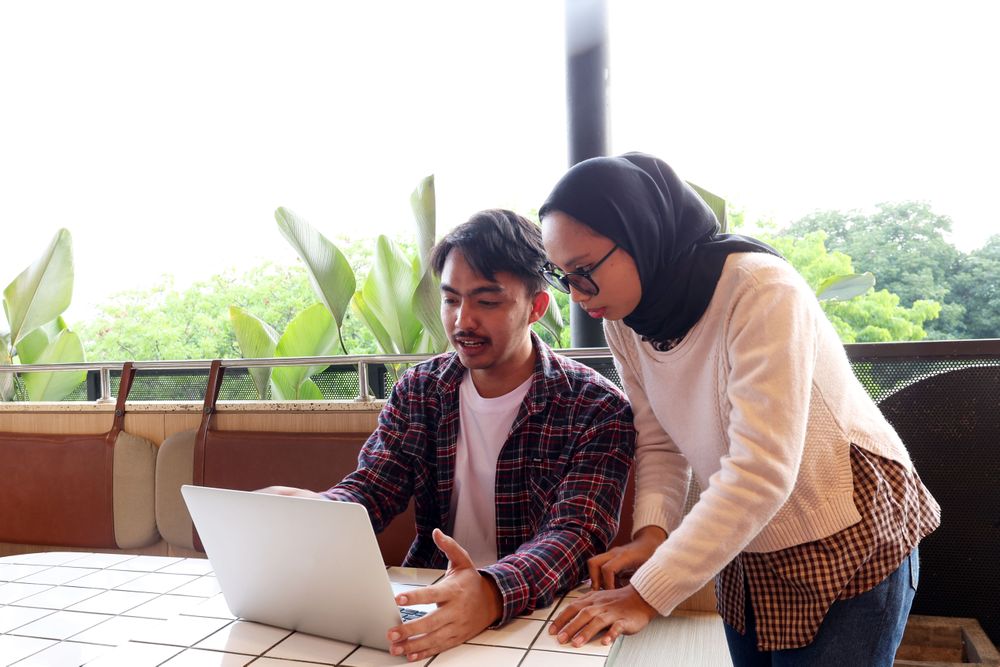 Two Young Asian Working People, Discussing at the Cafe Table, Working with Laptop Explaining File on Screen