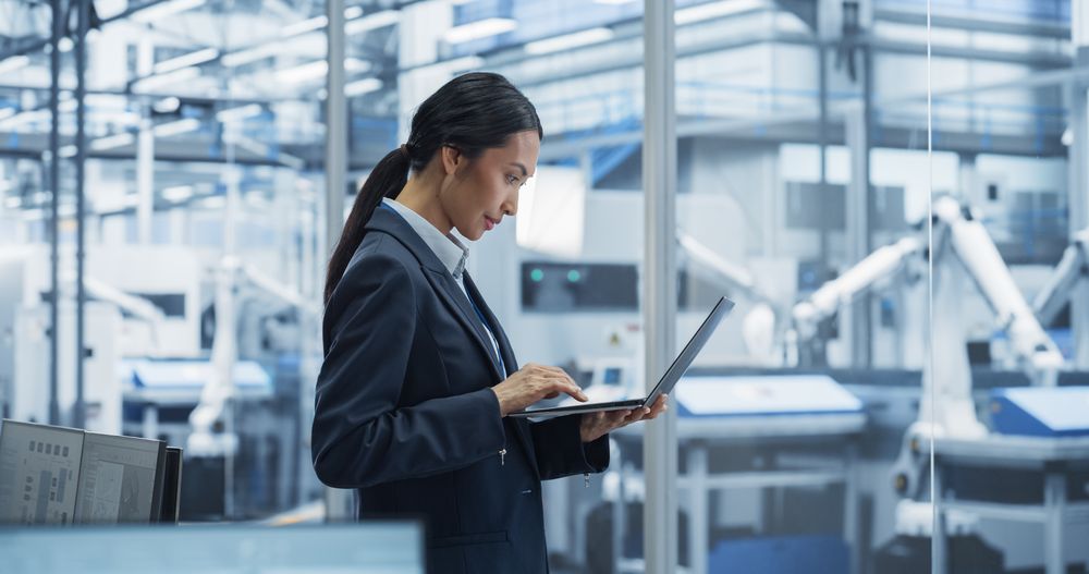 Production Line Technician Working on a Laptop Computer at a Factory Office. Asian Female Software Specialist Updating Code for Machine Production and Assembly with Automated Robotic Arms
