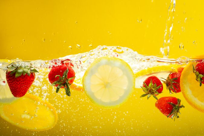 Orange & lemon slices with strawberries floating in water in yellow background