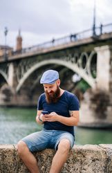 Bearded young man in casual wear using smartphone while walking on quay