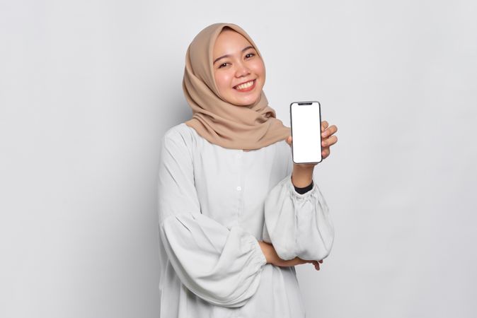 Confident Asian Muslim woman in a bright studio shoot presenting cell phone with mockup screen