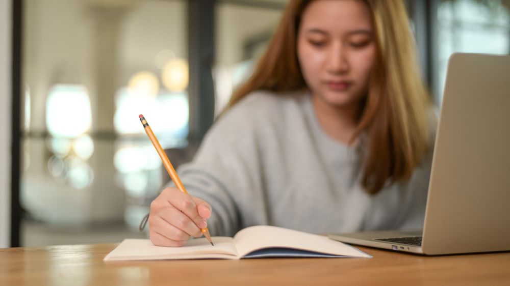 Close up shot of Asian woman writing on notebook with laptop on desk during online class, studying online at home.
