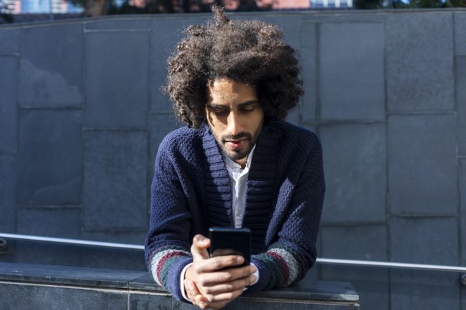 Black man in cardigan leaning forward outdoors on sunny day using cell phone