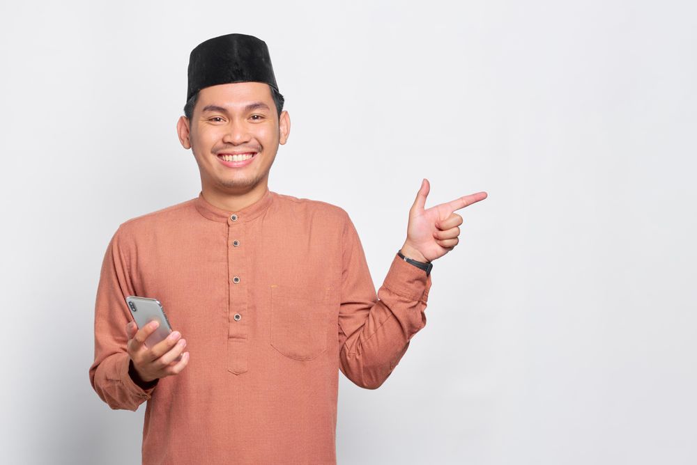 Happy Muslim man in kufi hat holding mobile phone and pointing to the side