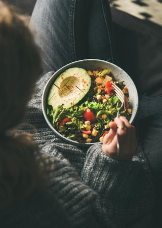 Woman in jeans and sweater eating bowl of fresh salad, top view