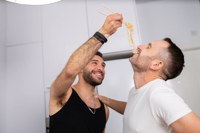 Laughing male couple playfully tasting pasta as they prepare meal