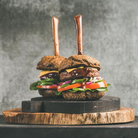 Two cheeseburgers with knives, stacked with fresh vegetables, on wooden board, square crop