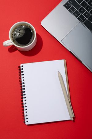 Top view of laptop keyboard with mockup screen on red table with coffee and notebook with copy space