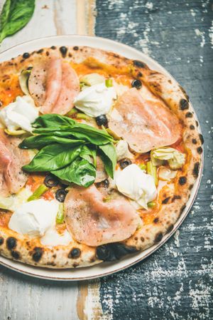 Close up of freshly made wood fire oven pizza with spinach, ham and olives, on wooden background