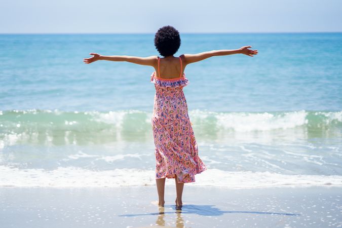 Rear shot of female in maxi dress with outstretched arms facing blue ocean