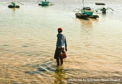 Back view of fisherman holding a wading bucket in water near boats bEJ2M0