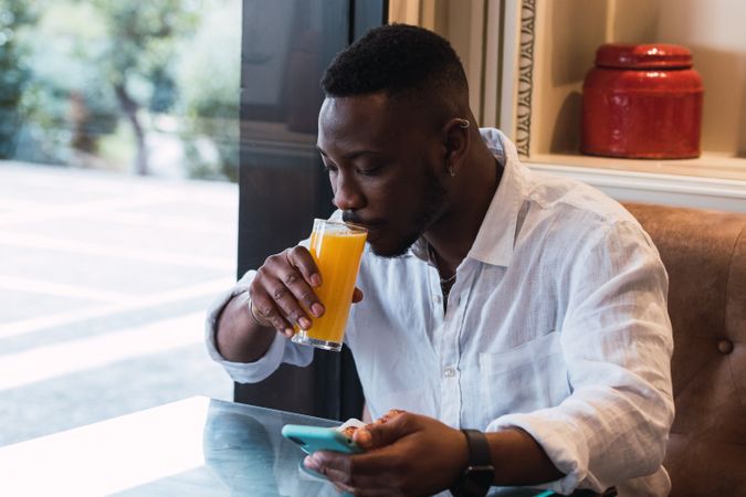 Black man browsing on phone in cafe while sipping an orange juice