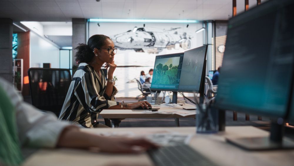 Black Female Game Developer Using Desktop Computer With 3D modelling Software And Talking to Asian Female Project Manager in Game Design Studio Office.