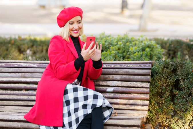 Female in red outfit and beret sitting on wooden bench in park checking phone