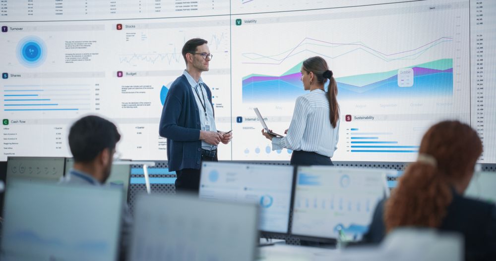 Male Data Scientist And Female Project Manager Discussing Reports And Graphs On Big Digital Screen In Modern Monitoring Office.