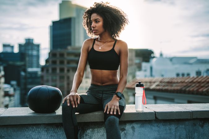 Athletic woman sitting on rooftop fence with a medicine ball and water bottle during workout
