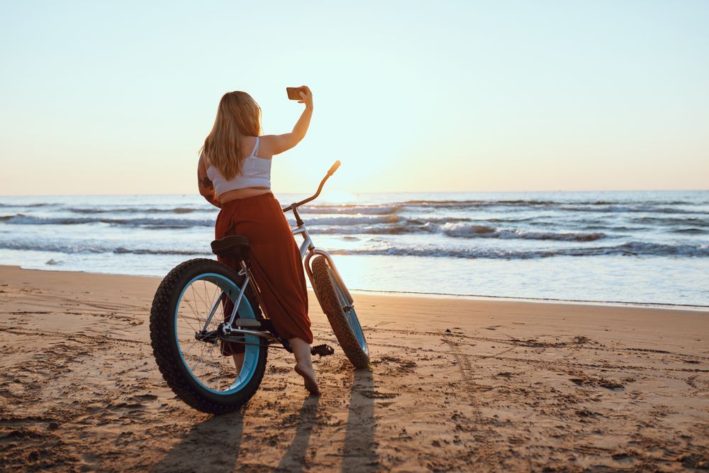 Cheerful woman taking selfie on smartphone while resting with bicycle at sunny shore