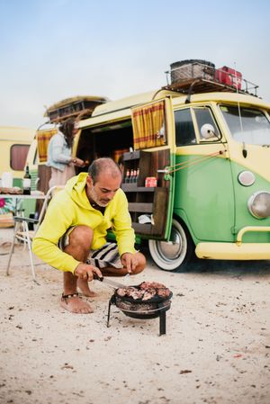 Tanned man cooking on camping stove beside parked mini van